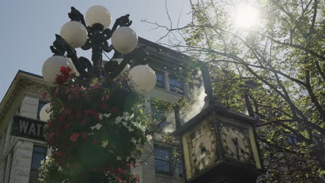 Reloj-De-Vapor-Del-Distrito-De-Vancouver-Gastown,-Con-Vapor-Saliendo,-Llamaradas-Solares-Y-Una-Pluma-Volando-A-Través-De-La-Imagen,-Y-Una-Farola-Con-Flores-Colgantes,-Cámara-Lenta-De-Mano