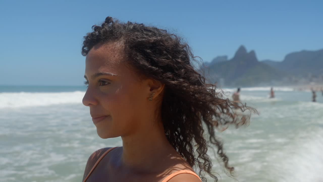 Bidikan tubuh bagian atas seorang wanita muda multiras tersenyum dan melihat ke kiri di pantai Ipanema. Angin menerbangkan rambutnya saat orang-orang menikmati ombak.