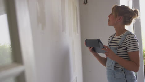 woman with digital tablet decorating room in house looking at tester paint colour strips on wall