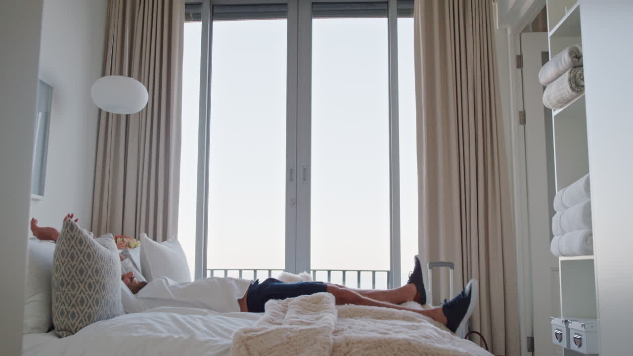 행복한 남자가 침대에 뛰어 성공적인 여행 여행 후 휴식을 취하고 미소 짓고 독립적인 생활 방식의 자유를 즐기고 있습니다.