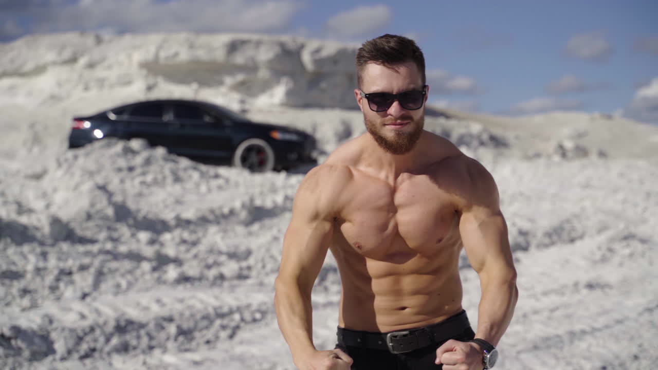 Athletic man posing on camera. Strong bodybuilder without shirt shows his trained body on white canyon background. Muscular sportsman outdoors.