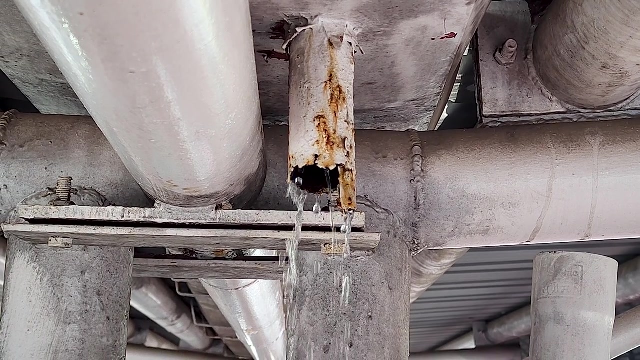 Rusten industrirør lækker vand fra korroderet samling under metalstruktur, hvilket viser infrastrukturskader, VVS-fejl og vedligeholdelsesproblemer.