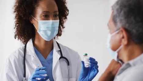 Mixed-race-female-doctor-wearing-mask-giving-vaccination-to-senior-man-at-home