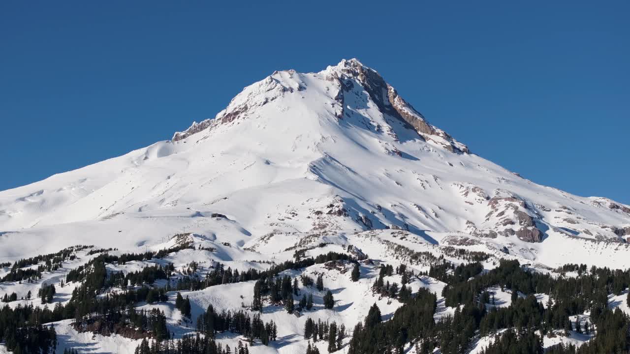 Una ripresa aerea con drone a media distanza del Monte Hood, dei suoi ghiacciai e della fitta coltre di neve sulla cima del vulcano isolato, di fronte a una pineta nel Pacific Northwest, vicino a Hood River e Portland, Oregon
