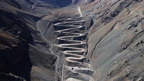 Dolly-out-aerial-view-of-the-cuesta-caracoles-road,-border-between-Chile-and-Argentina,-cargo-trucks-passing-through-the-curves,-sunny-day