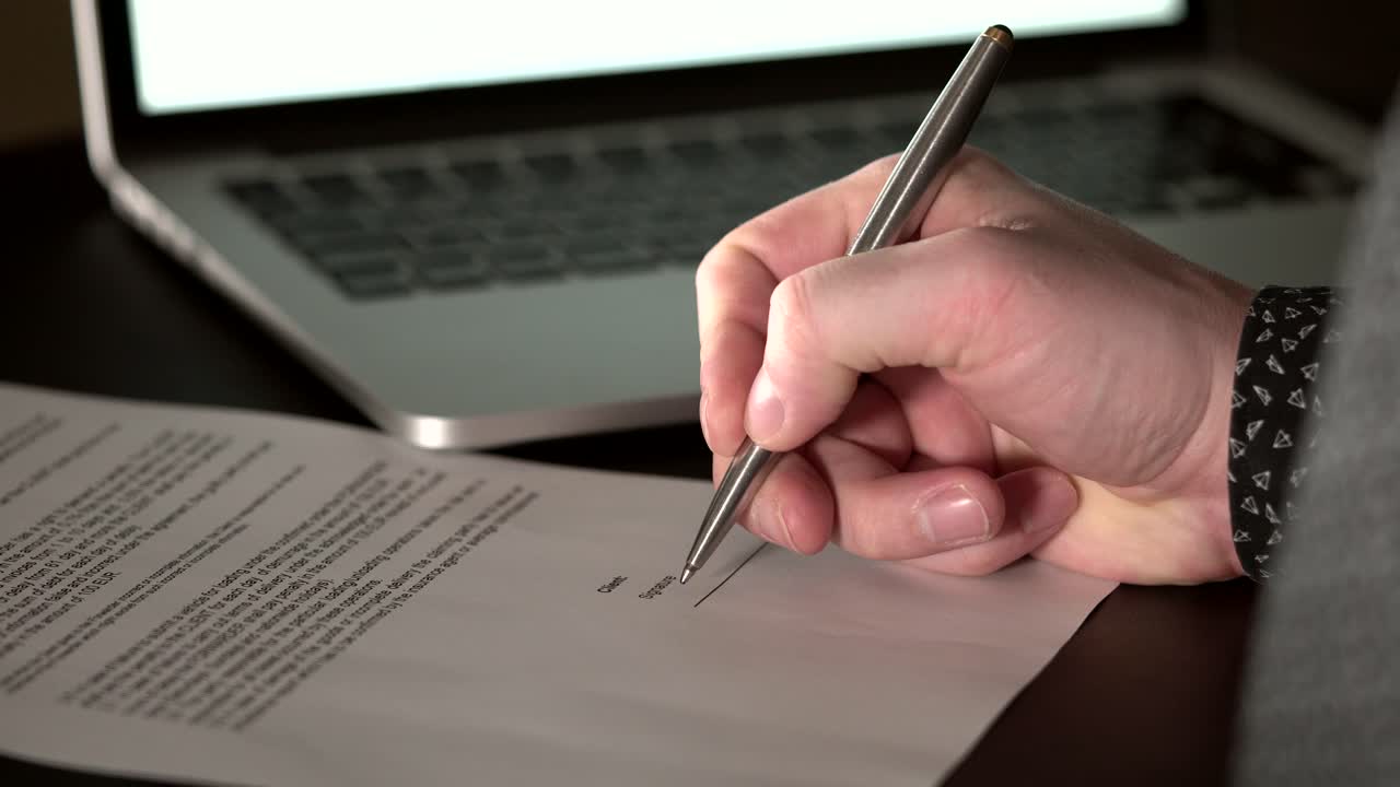 persona firmando un documento en un escritorio con una computadora portátil