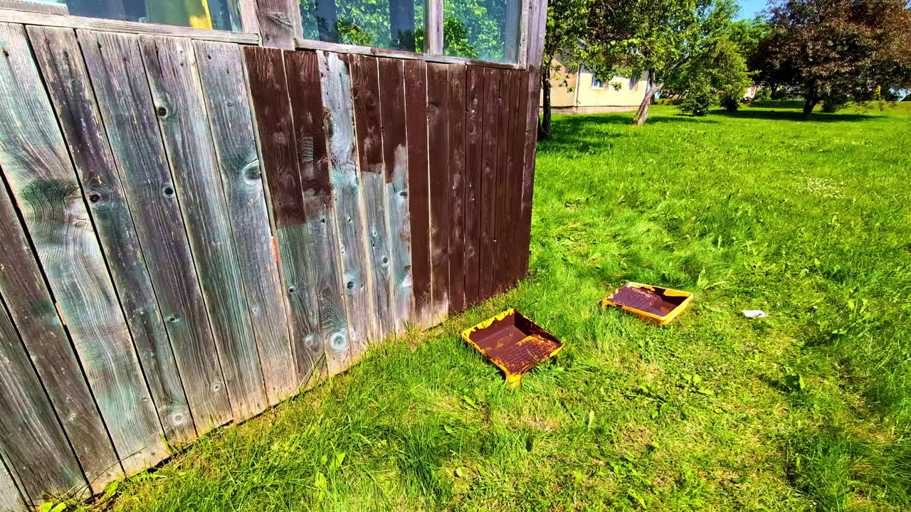 Paint Tray Lying On The Grass While Painting A Wooden Plank Wall Of A Structure. Close-up Shot