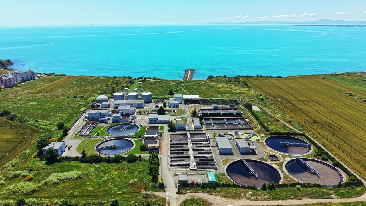 Water treatment facility located by the sea with large circular tanks and green fields visible in the background, taken on a clear day