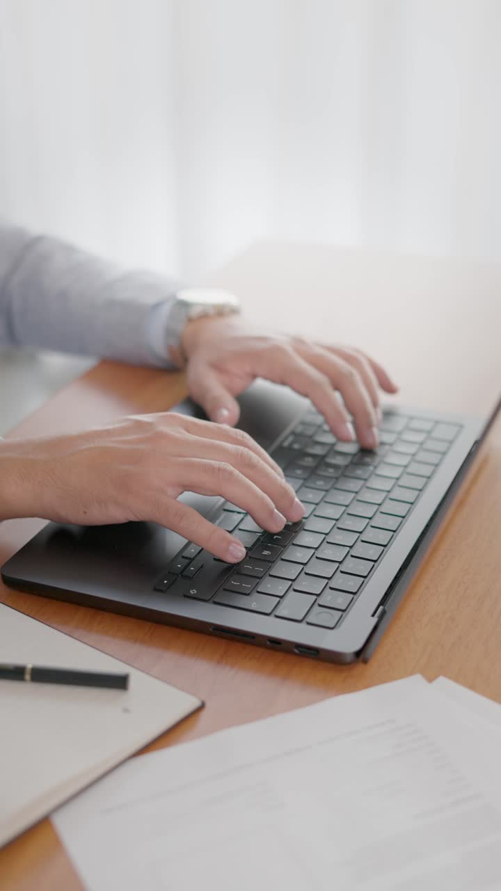 Inquadratura verticale di mani che digitano su una tastiera di un laptop con documenti e penna su una scrivania di legno
