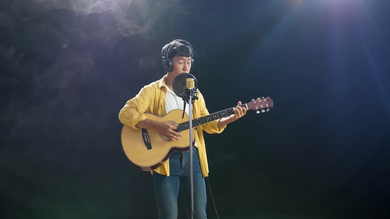 joven asiático con auriculares tocando una guitarra y cantando en un micrófono condensador sobre el fondo blanco de humo negro