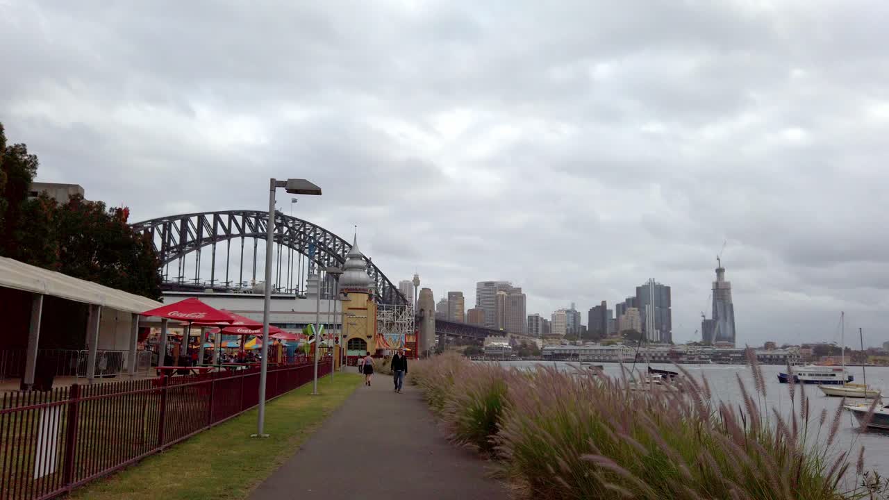 Waterfront walking shot with Harbour Bridge, fast food restaurant and downtown buildings in the distance on an overcast day, walking forward with pan right shot