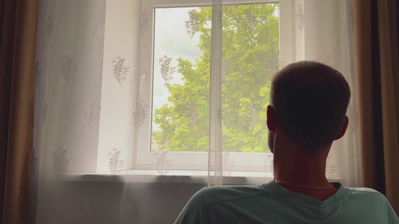 Eine Rückenansicht eines Mannes in einem Zimmer, der durch ein Fenster mit Spitzenvorhängen auf grüne Bäume draußen blickt. Geeignet für Anzeigen zu psychischer Gesundheit, Immobilien oder Lifestyle.