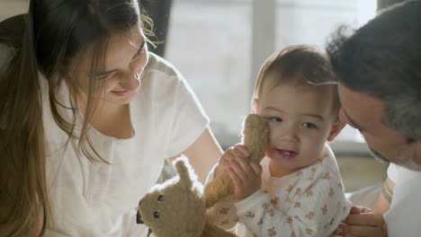 padres felices en la cama el domingo por la mañana jugando con su linda niña que sostiene un oso de peluche