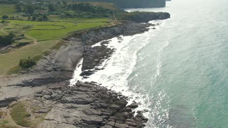 a drone rises slowly, over the sea as the waves crash relentlessly against jagged grey rocky shoreline, revealing endless lush fields of green