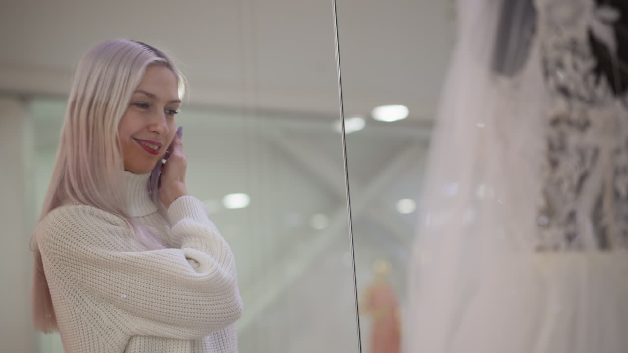 bride to be smiles while browsing wedding gown display behind glass window in mall boutique, mannequins reflected in soft light as lace details shimmer in elegant showroom setting