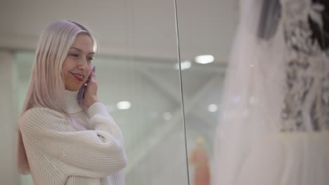bride to be smiles while browsing wedding gown display behind glass window in mall boutique, mannequins reflected in soft light as lace details shimmer in elegant showroom setting