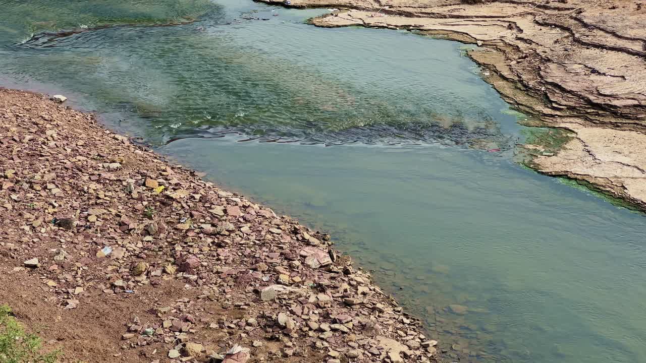 A close static shot showing a shallow river gently flowing between rocky banks, with clear greenish-blue water revealing stones beneath, capturing the raw beauty of a natural rural water stream