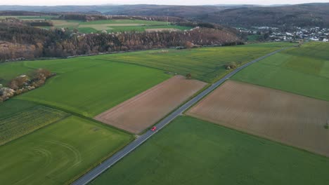 A-red-car-driving-along-a-country-road-that-leads-through-green-and-brown-fields-and-meadows