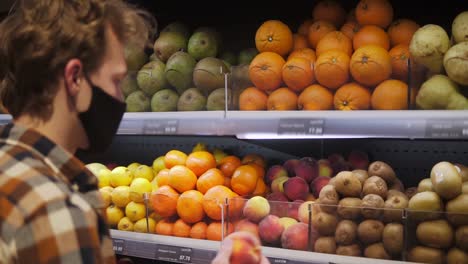 giovane in camicia a quadri, indossando un respiratore, una maschera nera di tessuto che compra frutta con un sacchetto di plastica nel supermercato durante la pandemia di quarantena covid-19 coronavirus