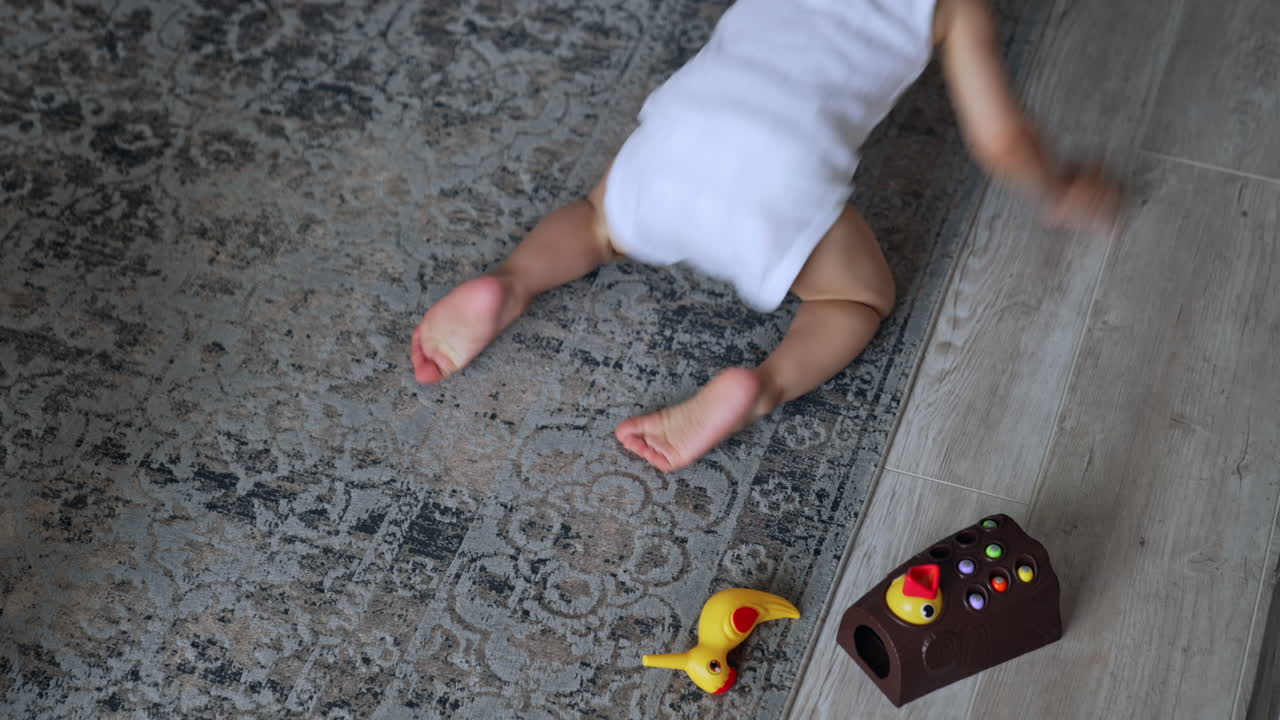 Little adorable baby boy sits on the carpet. Kid holds a toy looking at camera and then crawls away. Top view.