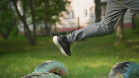 close-up view of a person wearing gray trousers and sneakers jumping over tires placed on a grassy field in an outdoor exercise, with trees and a building blurred in the background