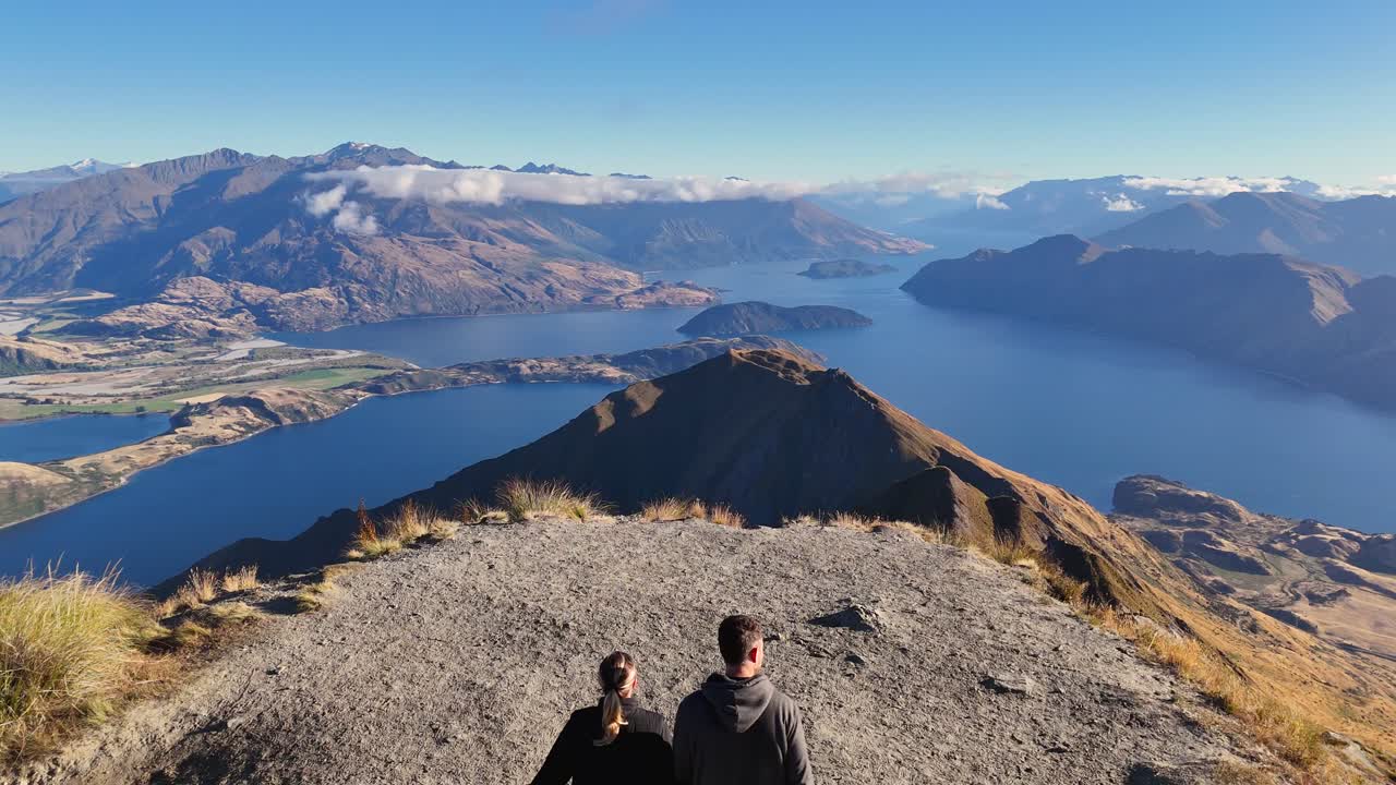 A 4K drone follows a couple walking up to the iconic Roy’s Peak viewpoint in Wanaka, New Zealand. Scenic mountains, lakes, and alpine trails surround the stunning hike