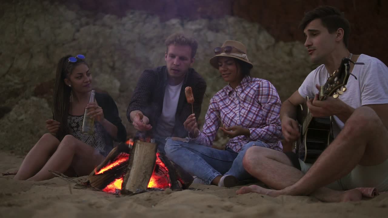 пикник молодых людей с кострем на пляже вечером. веселые друзья поют песни и играют на гитаре. замедленное движение