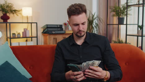 smiling happy young guy at home counting money cash use smartphone, income, saves, lottery win