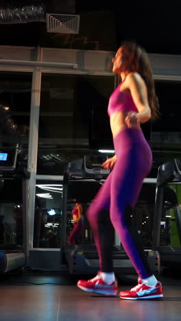 side view of a full-length woman in sportswear jumping on a rope in gym. long-haired sportswoman in pink top and leggings training on a skipping rope on the background of fitness center. vertical video