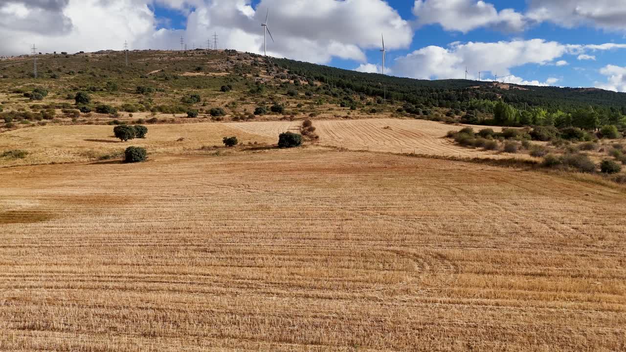 A low altitude Dolly Forward shot glides rapidly over a dry golden field towards a ridge of wind turbines generating energy in the rural landscape of Segovia, Spain
