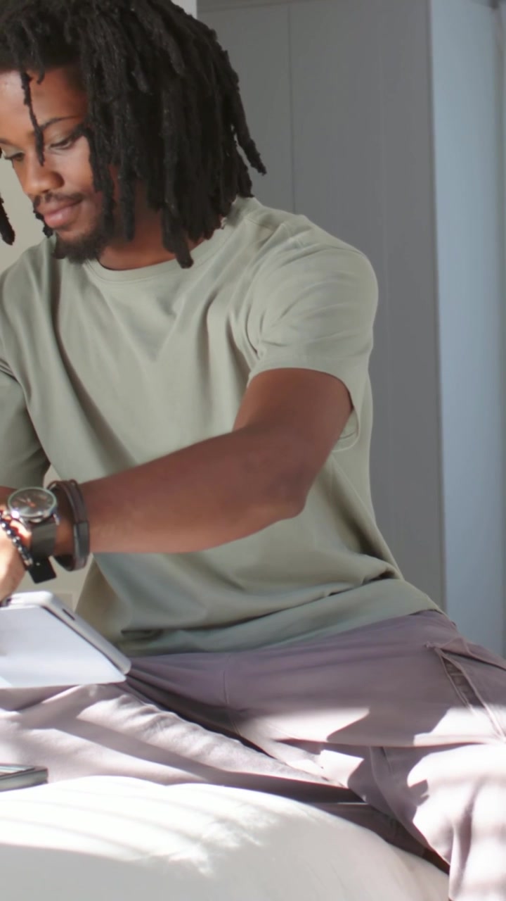 Vertical video: Man wearing green tee picking up white tablet and arranging devices on bed for work