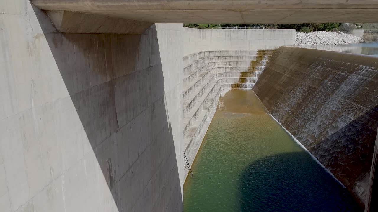 Water cascading over a large concrete dam, highlighting its structure, the green water below, and the natural surroundings during daytime hours