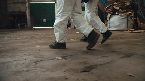 Close-up-shot-of-two-men-in-white-uniforms-and-black-shoes-walking-along-the-huge-hall-of-a-waste-processing-plant.-Two-male-colleagues-walk-with-their-feet-on-the-floor-in-a-large-waste-recycling-plant