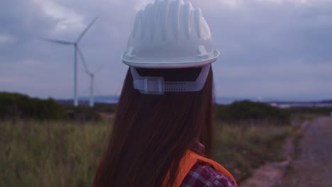 rear closeup of woman technician wear protective helmet looks at wind turbines