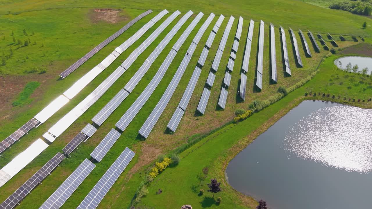 Aerial view of a large solar farm with rows of photovoltaic panels near a pond, surrounded by green fields, producing sustainable, renewable energy, parallax shot