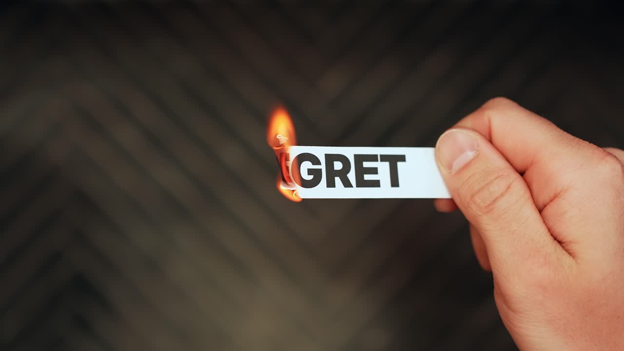 Close-up of a hand holding a paper strip labeled “REGRET” as the edge burns with flame on a dark background. Letting go of past mistakes and emotional healing concept