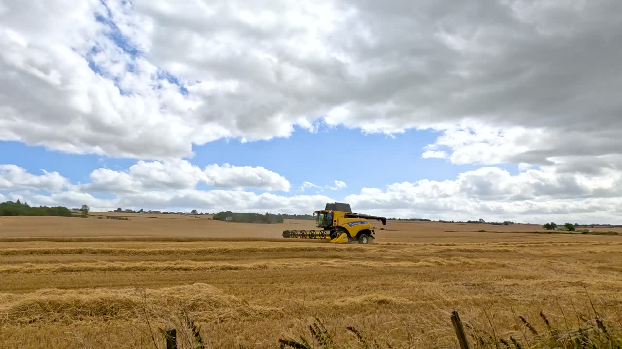 A yellow combine harvester moves steadily across a golden wheat field in Cromarty, Scotland, under partly cloudy daylight, captured in a wide static shot