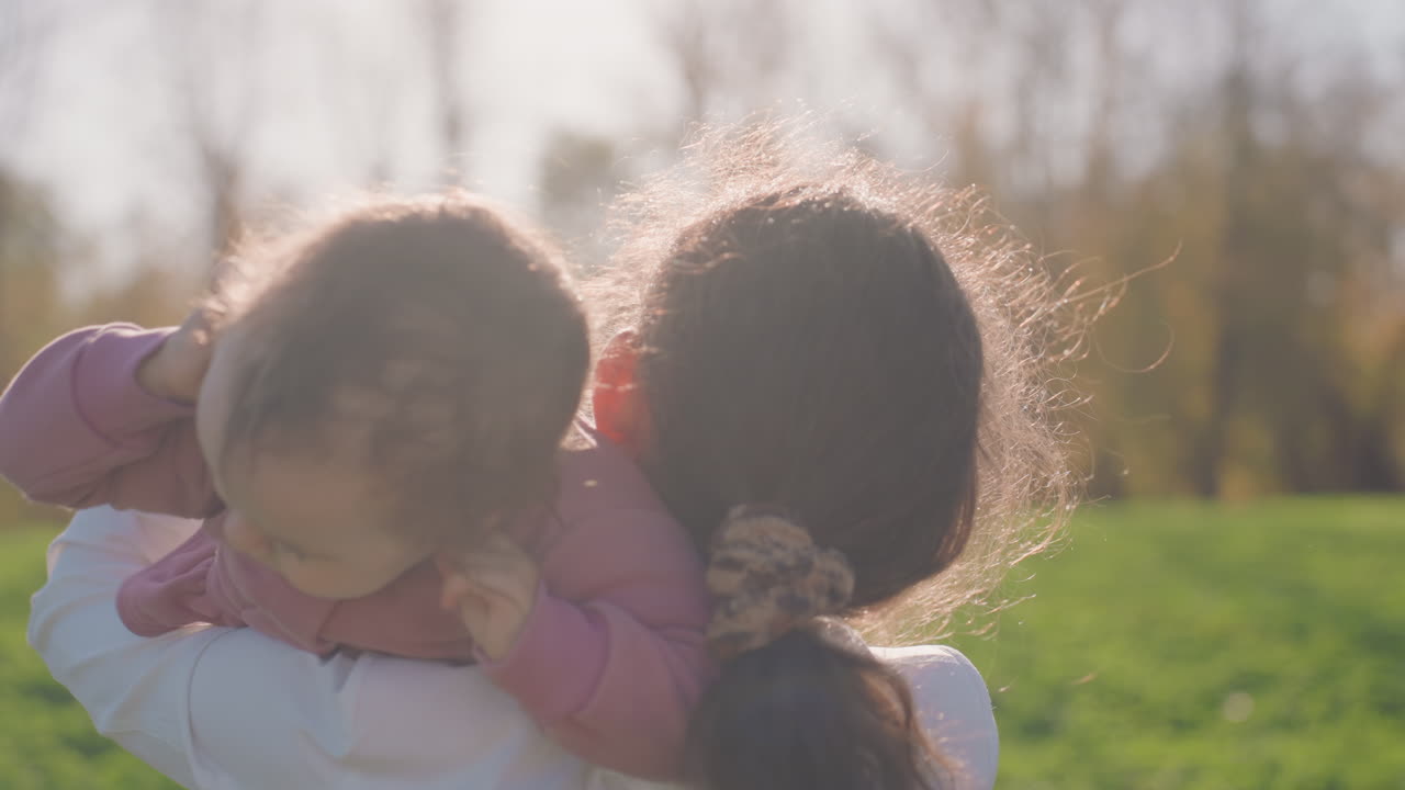Un genitore solleva il bambino alla luce del sole, in un abbraccio intimo e un dondolio delicato, con giacca rosa e cappello bianco su sfondo di un parco autunnale, un morbido alone di capelli dalla retroilluminazione, occhi teneri e manine che si stringono.