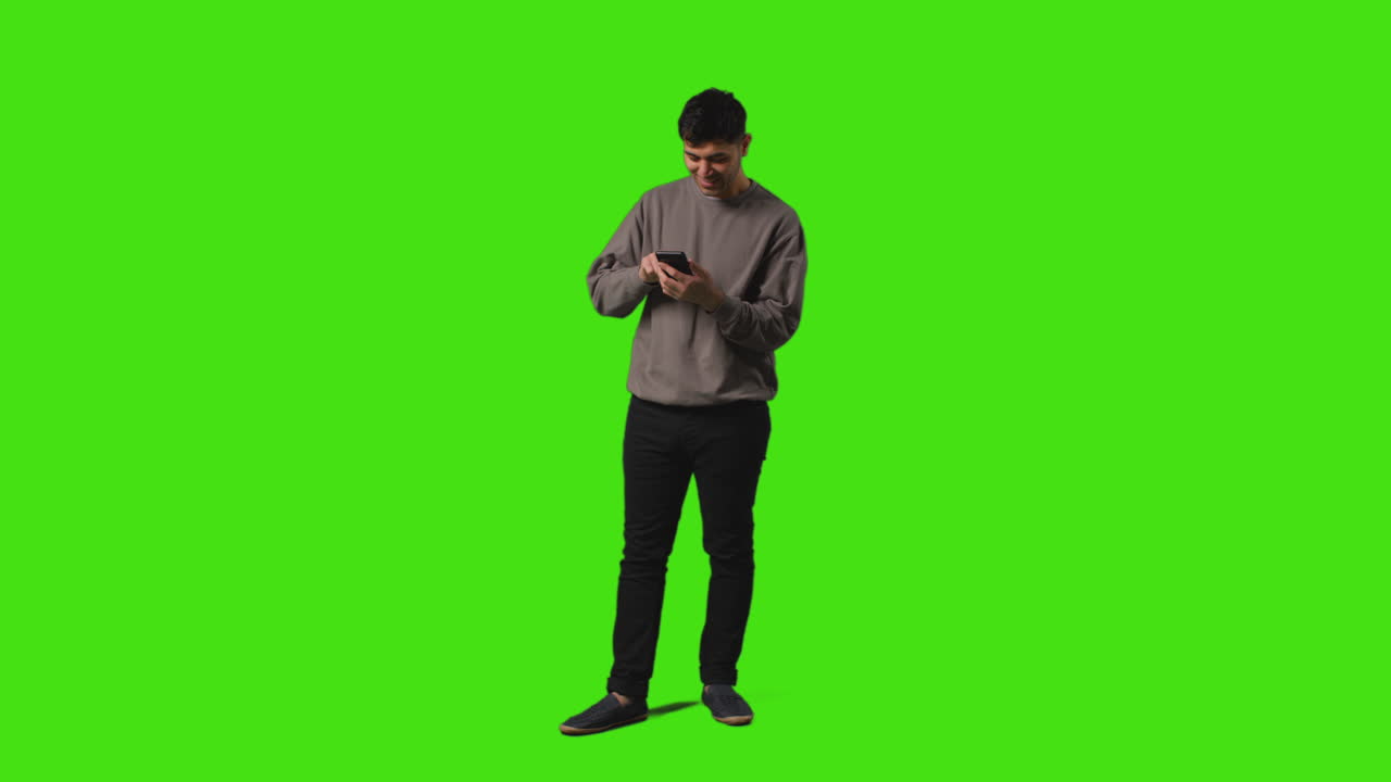 Young Man Sending Or Reading Text Message Or Scrolling Online On Mobile Phone Standing Against Green Screen 1