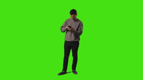 Young-Man-Sending-Or-Reading-Text-Message-Or-Scrolling-Online-On-Mobile-Phone-Standing-Against-Green-Screen-1