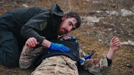 nahaufnahme eines selbstbewussten männlichen militärischen brünetten mit bart in einer dunkelgrünen militäruniform hört auf die atmung und hält die brust eines verwundeten soldaten während des kampfes und bietet erste hilfe außerhalb der stadt