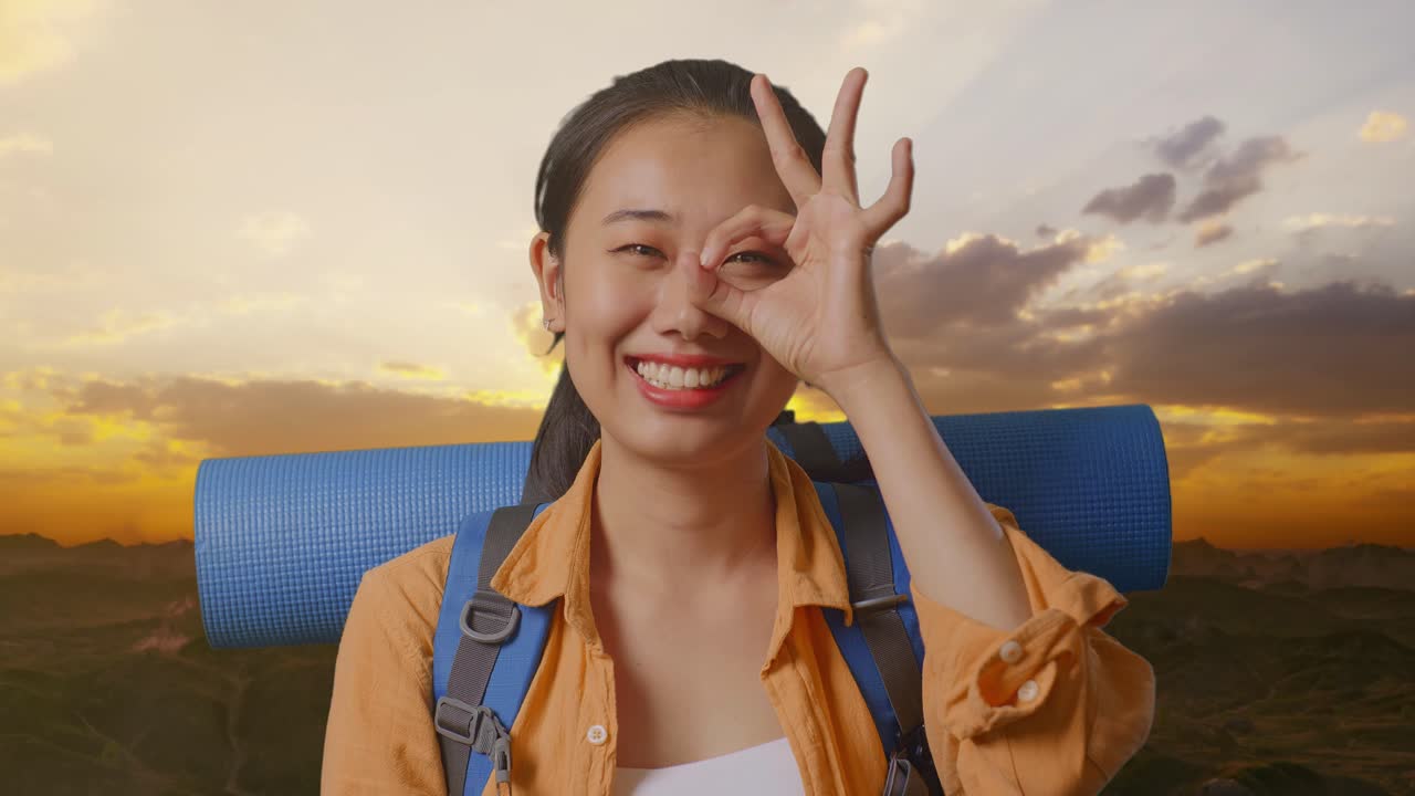 Close Up Of Asian Female Hiker With Mountaineering Backpack Smiling And Showing Ok Sign With Fingers Over Eyes While Standing On The Top Of Mountain During Sunset Time