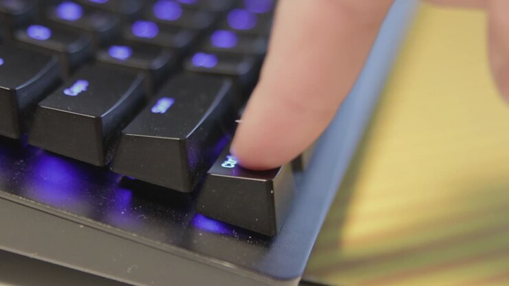 Hand pressing a single key on a slightly dusty, black keyboard with blue backlit keys