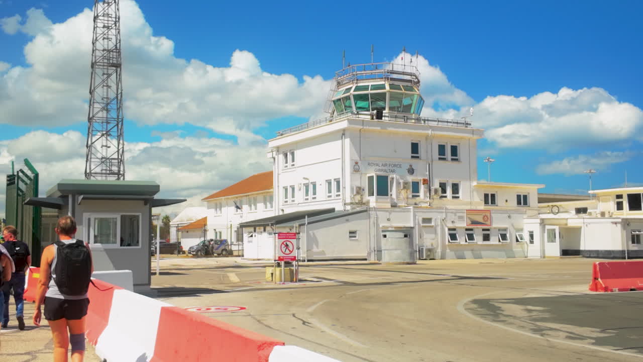 People walking towards an air traffic control tower under a partly cloudy sky, showcasing a travel scene at a small airport - Gibraltar