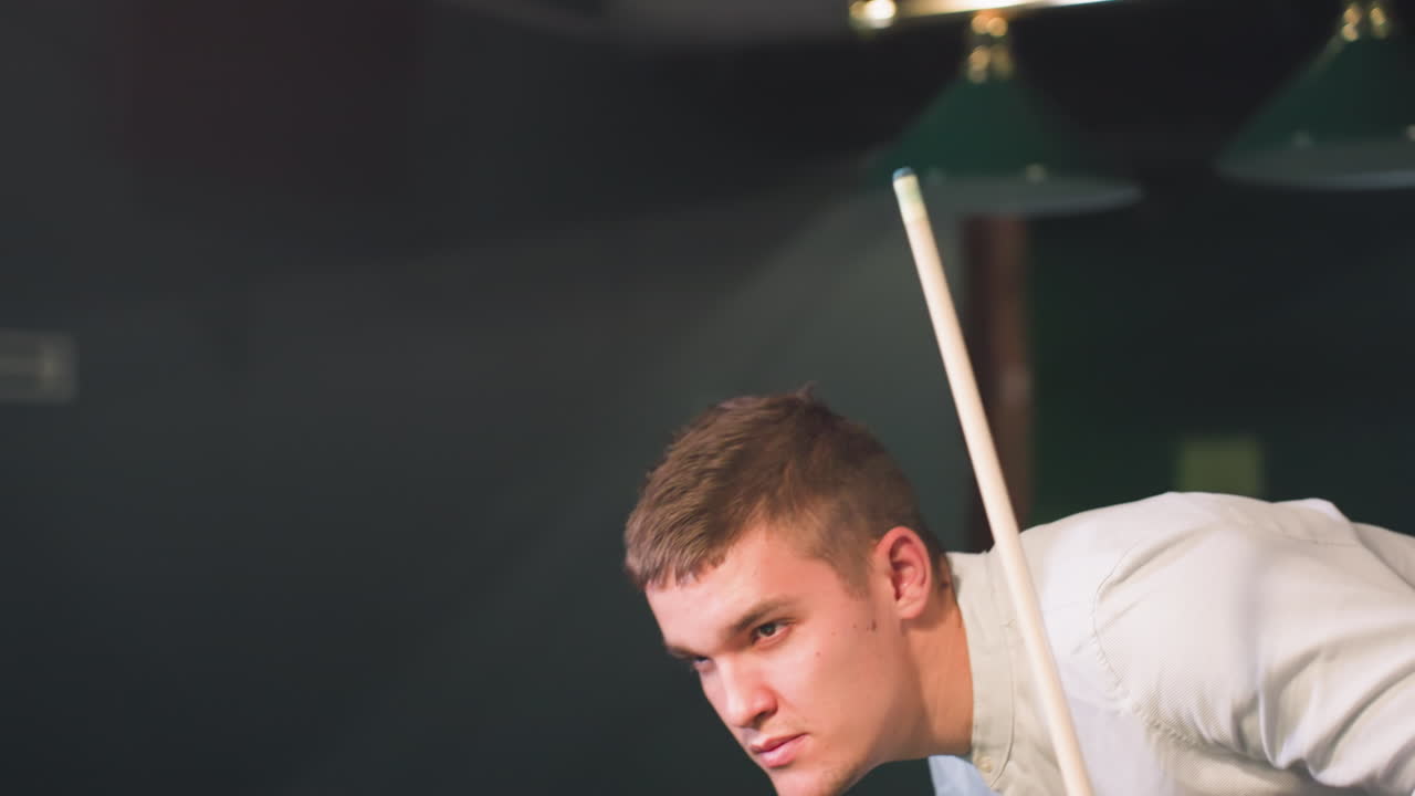 차분한 표정으로  셔츠를 입은 젊은 남자의 클로즈업, 큐 스틱과 레몬 음료를 들고 빌리어드 홀에서 걸어가는 동안. 오버헤드 조명은 부드러운 빛을 내며, 세련되고 편안한 분위기를 향상시니다.