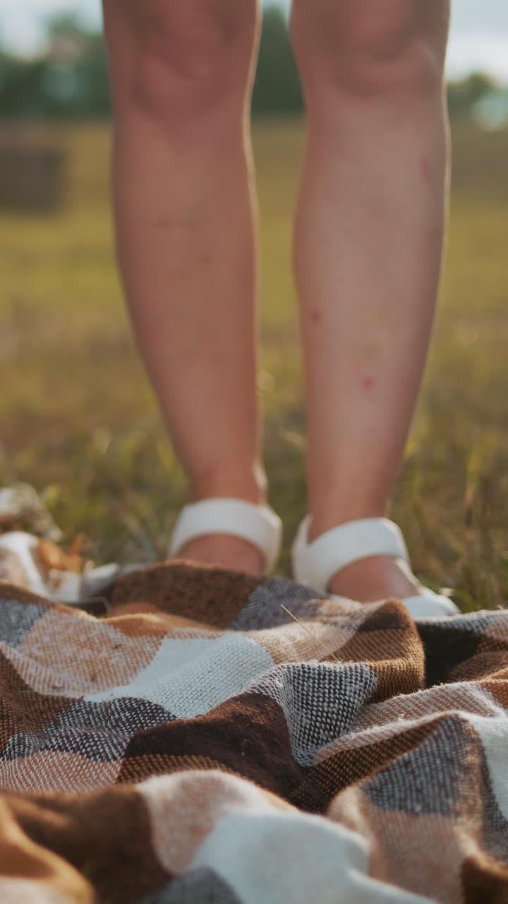 Close-up of checkered picnic blanket spread on grassy field with woman in white sandals gently lowering a woven basket filled with snacks, warm countryside atmosphere with hay bale