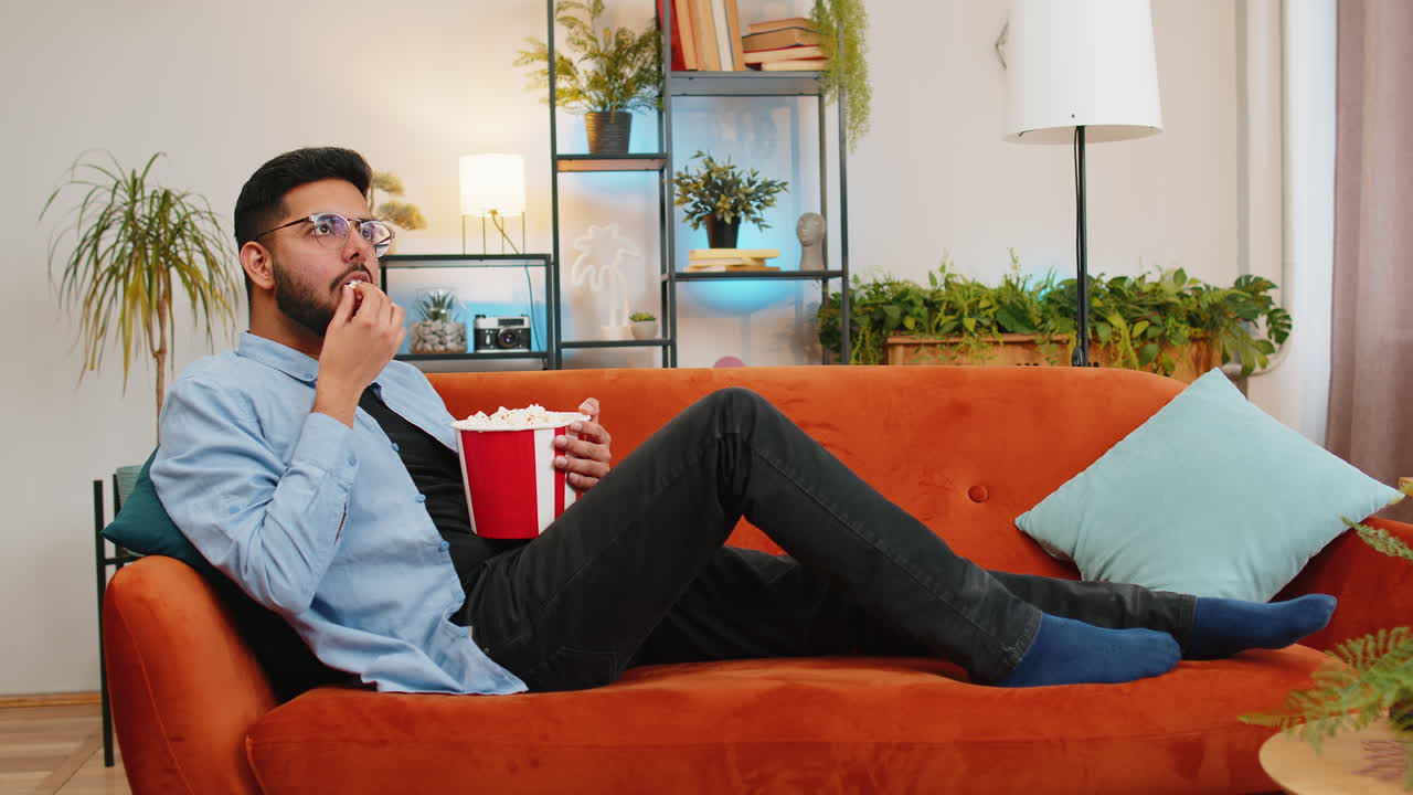 hombre indio relajado comiendo palomitas de maíz y viendo películas disfrutando de películas durante el fin de semana acostado en el sofá de casa