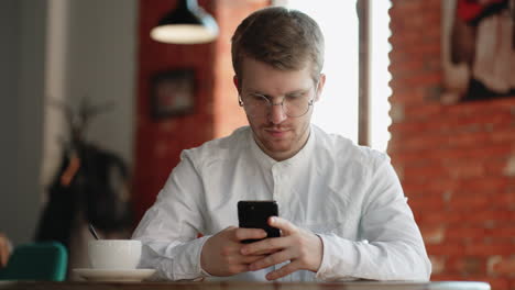 handsome man in white shirt and with glasses is sitting alone in coffee shop surfing internet by smartphone