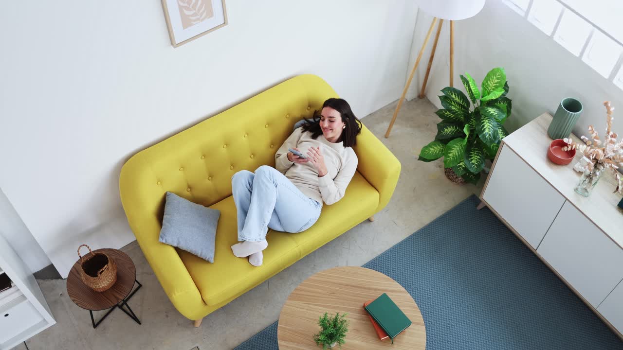 Ung kvinde slapper af på sofaen og bruger smartphone i stuen