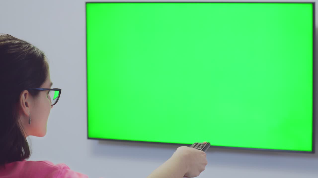 Brunette girl in glasses watching TV sitting in front of the TV, switching channels with the remote control rear view, chroma key